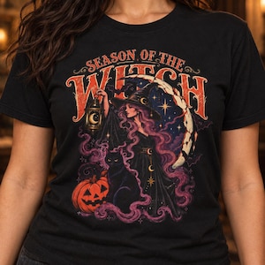 May include: A black t-shirt featuring a graphic print with the text "SEASON OF THE WITCH" in orange. The design depicts a witch in a pointed hat holding a lantern, a black cat, a jack-o'-lantern, bats, and a crescent moon amidst purple smoke.
