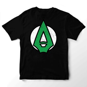 Może przedstawiać: Czarny t-shirt z krótkim rękawem z centralną grafiką przedstawiającą zielony grot strzały z czarnym obrysem na białym, okrągłym tle. Zielony wzór grota strzały ma widoczną, postarzaną i przetartą teksturę na całej powierzchni.