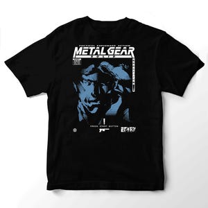 May include: A black t-shirt featuring a blue and white graphic of a character face. The text reads TACTICAL ESPIONAGE ACTION METAL GEAR SOLID, PRESS START BUTTON, and includes a small gun icon, a price tag of 99.99, and Japanese characters.