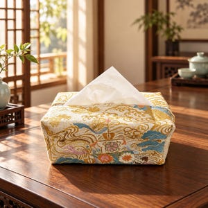 May include: A rectangular tissue box cover featuring a woven fabric with a gold, blue, and pink floral and wave pattern. A single white tissue is pulled through the top opening, standing upright in a triangular shape against the patterned textile.