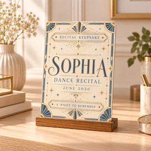 May include: A rectangular wooden plaque with a cream-colored surface featuring blue and gold decorative borders. The text reads: RECITAL KEEPSAKE, SOPHIA, DANCE RECITAL, JUNE 2026, A NIGHT TO REMEMBER. The plaque is supported by a dark wood base.