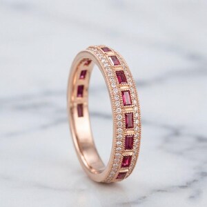 Peut inclure: Une bague en or rose ornée d'une rangée centrale de pierres rectangulaires rouge foncé taille baguette, encadrée par deux rangées extérieures de petites pierres rondes transparentes serties pavé. Les bords présentent un grain fin.