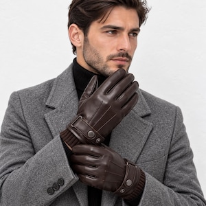 May include: Dark brown leather gloves featuring a ribbed knit cuff, a decorative strap with a metallic snap button closure, and vertical stitched detailing on the back of the hand. The gloves are worn with a grey wool coat and a black turtleneck.