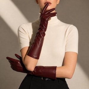 Puede incluir: Un par de guantes largos de cuero color burdeos intenso que se extienden más allá del codo. Los guantes presentan un acabado liso y mate con costuras visibles a lo largo de los dedos y el dorso de la mano, combinados con un top de cuello alto.