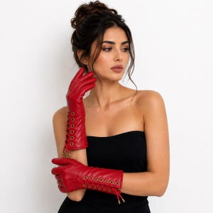 May include: A pair of vibrant red leather gloves featuring a long cuff design with gold-toned metal eyelets and matching red lace-up detailing that runs along the outer forearm, providing a secure fit and a distinct visual texture to the accessory.