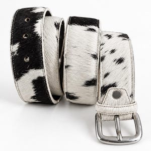 Puede incluir: Un cinturón con estampado de piel de vaca en blanco y negro y un acabado texturizado de pelo. El cinturón incluye una hebilla de metal plateado, una trabilla de cuero a juego y tres orificios circulares perforados para ajustar la talla.