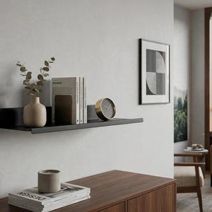 Puede incluir: Un estante de pared flotante negro mate que sostiene un jarrón de cerámica beige con tallos de eucalipto, tres libros con lomos blancos y un reloj analógico circular con borde dorado y esfera oscura, montado en una pared gris claro.