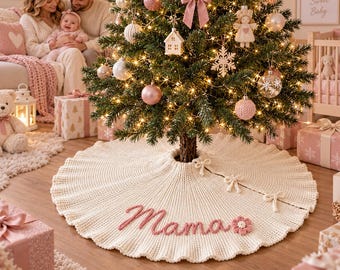 May include: A cream-colored, circular, ribbed knit Christmas tree skirt featuring the word "Mama" embroidered in pink yarn script, accented with a small pink flower applique and three matching ribbon ties along the side slit.