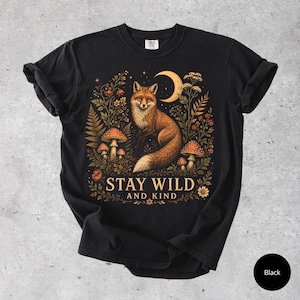 Könnte beinhalten: Ein schwarzes Kurzarm-T-Shirt mit einem Grafikdruck eines Fuchses, umgeben von Pilzen, Farnen und einer Mondsichel. Der Text "STAY WILD AND KIND" ist in hellbeiger Schrift unter der Fuchsillustration auf der Vorderseite des Shirts gedruckt.
