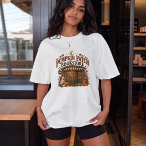 Puede incluir: Una camiseta blanca de manga corta con un estampado gráfico de una tienda y el texto PUMPKIN PATCH BOOKSTORE. El diseño incluye hojas de otoño, calabazas, una luna creciente y una pequeña figura de fantasma en tonos marrones y naranjas.