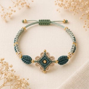May include: A macrame bracelet featuring a central flower-shaped motif in sage green and cream cord, accented with small gold-toned beads. The band consists of braided sage green cord with additional leaf-shaped elements and a sliding knot closure.