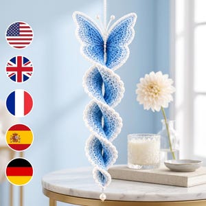 Puede incluir: Un adorno de mariposa de ganchillo colgante con un diseño de cola en espiral en tonos azules y blancos. La mariposa tiene un cuerpo azul detallado con borde blanco, dos antenas con pequeñas cuentas blancas en las puntas y una cuenta blanca abajo.
