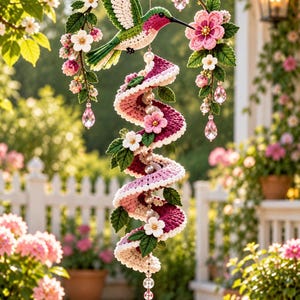 May include: A crochet wind spinner featuring a green and pink hummingbird perched on a branch with white flowers, connected to a spiraling pink and white crochet tail adorned with small flowers, green leaves, and a dangling pink crystal teardrop.