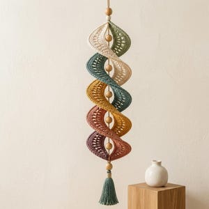 Könnte beinhalten: Ein hängendes Spiral-Mobile mit gehäkelten Stoffsegmenten in Creme, Salbeigrün, Petrol, Senfgelb, Terrakotta und Pflaume. Die Segmente sind durch glatte Holzperlen an einer zentralen Schnur getrennt und enden mit einer salbeigrünen Garnquaste.