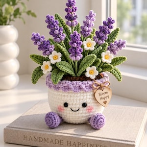 Op de afbeelding: Een gehaakte plant in een pot met paarse lavendelstengels en kleine witte bloemen met gele harten. De witte pot heeft een lachend gezicht met roze wangen, paarse voetjes en een houten hartvormig label met de tekst Bloom Happy.