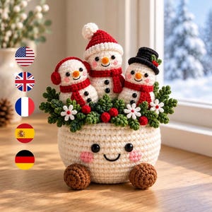 May include: A crochet display featuring three small snowmen with orange noses and red scarves nestled in a cream-coloured bowl with a smiling face. The snowmen wear a red hat, red earmuffs, and a black hat, surrounded by green leaves and white flowers.