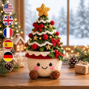 Op de afbeelding: Een gehaakte kerstboomdecoratie in een crèmekleurige pot met een lachend gezicht, roze wangen en bruine voetjes. De groene boom is versierd met rode strikken, kleine gouden bolletjes, gele sterren en een grote gele ster als piek.