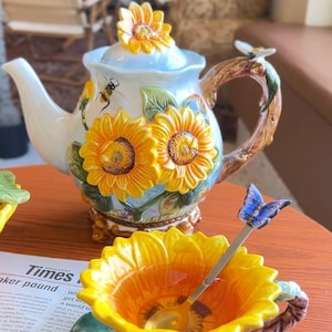 Può includere: Una teiera e una ciotola in ceramica abbinate con disegni di girasoli gialli in rilievo, foglie verdi e api dipinte. Il coperchio della teiera ha un pomello a forma di girasole e il manico ricorda un ramo marrone con una piccola ape. Un cucchiaio argentato con una farfalla blu poggia nella ciotola.