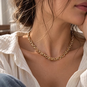 May include: A gold-toned chain necklace featuring a series of interlocking, textured oval links. The necklace rests against the neck, showcasing a polished metallic finish and a consistent, repeating pattern throughout the entire length of the chain.