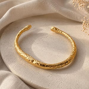 May include: A gold-toned cuff bracelet featuring an irregular, hammered texture across its entire surface. The bracelet has an open-ended design with rounded terminals, showcasing a reflective metallic finish and a distinct, organic, uneven shape.