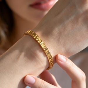 May include: A gold-toned bangle bracelet featuring a distinctively hammered, uneven surface texture. The metal band has a reflective finish that catches the light, creating highlights and shadows across its irregular, organic-looking exterior.