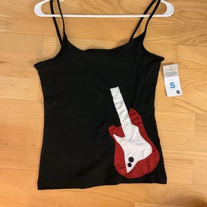 Könnte beinhalten: Ein schwarzes Trägertop mit einer rot-weißen Stoffapplikation in Form einer Gitarre auf der unteren rechten Seite. Das Gitarrendesign enthält zwei kleine schwarze runde Knöpfe. Ein weißer Kunststoffkleiderbügel ist oben befestigt und ein S-Etikett.