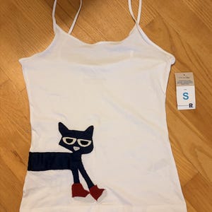 Könnte beinhalten: Ein weißes Trägershirt mit dünnen Spaghettiträgern und einer dunkelblauen Katzen-Applikation mit weißer Sonnenbrille und roten Schuhen. Ein weißes Verkaufsetikett ist seitlich angebracht und zeigt die Größe S sowie den Buchstaben R in Fettdruck.