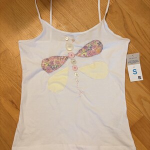 Puede incluir: Una camiseta de tirantes blanca con un gráfico de libélula hecho de botones y parches de tela. El cuerpo de la libélula consiste en una fila vertical de botones, mientras que las alas están hechas de tela floral y amarilla. Tiene una etiqueta de talla S.