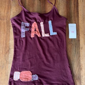 Op de afbeelding: Een donkerbordeauxrood hemdje met spaghettibandjes en het woord FALL in grote, veelkleurige stoffen letters op de borst. De letters zijn gemaakt van verschillende stoffen met patronen. Een klein, rechthoekig wit label met XS is aan de zijkant bevestigd.
