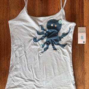 May include: A light blue camisole featuring a dark blue octopus appliqué on the front. The octopus is decorated with several small, round, white and light green buttons. A white retail tag with the letter S is attached to the side of the garment.