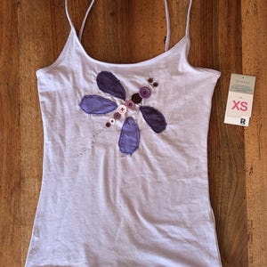 May include: A light purple spaghetti strap tank top featuring a dragonfly appliqué made of purple fabric patches and various brown and purple buttons. A white retail tag attached to the side displays the text "PRIMARK" and the size "XS".