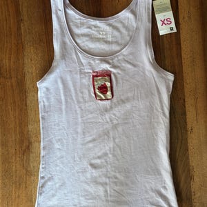 Peut inclure: Un débardeur sans manches lilas clair avec une encolure ronde. Le centre de la poitrine présente un petit écusson rectangulaire représentant un pot de confiture à la fraise rouge avec un couvercle à carreaux. Une étiquette blanche taille XS est fixée.