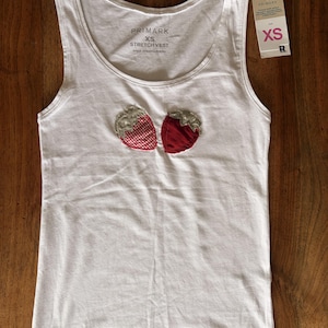 Può includere: Una canotta bianca elasticizzata senza maniche con scollo rotondo, caratterizzata da due applicazioni in tessuto a forma di fragola sul petto. Una fragola ha un motivo a quadretti rossi e bianchi, l'altra è rosso tinta unita. Etichetta taglia XS.
