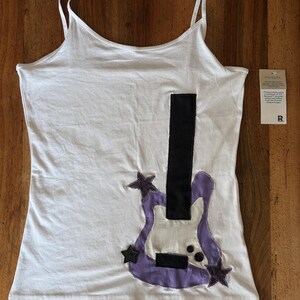 May include: A white spaghetti strap tank top featuring a large, purple and white electric guitar appliqué on the front. The guitar design includes a black neck, two black buttons, and three dark gray star-shaped accents. A product tag is attached.