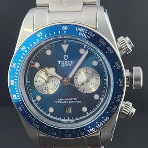 May include: A silver-tone stainless steel chronograph watch with a blue dial and a blue tachymeter bezel. The watch features the Tudor Geneve logo, two silver sub-dials, a date window at the six o'clock position, and a metal link bracelet.