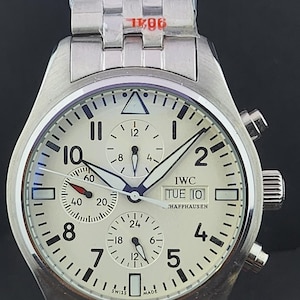 May include: A silver-tone stainless steel chronograph wristwatch with a cream-coloured dial, featuring black Arabic numerals, three sub-dials, a day-date window at the three o'clock position, and the text IWC SCHAFFHAUSEN and SWISS MADE.