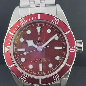 Könnte beinhalten: Eine Armbanduhr aus Edelstahl mit bordeauxrotem Zifferblatt und passender bordeauxroter Drehlünette. Das Zifferblatt zeigt weiße, leuchtende Stundenmarkierungen, das Tudor-Logo und den Schriftzug Tudor Geneve, 200m:660ft, Master Chronometer, Swiss Made.