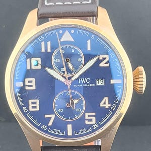 May include: An IWC Schaffhausen wristwatch with a bronze-toned case and a deep blue dial. The watch features large Arabic numerals, two sub-dials, a date window at the three o'clock position, and a dark brown leather strap with silver-toned rivets.