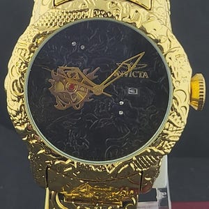 Puede incluir: Un reloj analógico de tono dorado con una correa y caja de metal con un intrincado patrón de dragón. La esfera negra muestra el nombre de la marca Invicta, manecillas doradas, una ventana de fecha a las tres y un motivo de dragón.