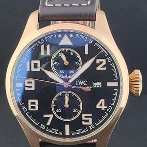 May include: A bronze-toned wristwatch with a dark blue dial, featuring large Arabic numerals, two sub-dials, and a date window at the three o'clock position. The watch has a brown leather strap with white stitching and two silver-toned rivets.