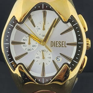 Op de afbeelding: Een goudkleurig polshorloge met een zilveren wijzerplaat met zonnestraalpatroon, het Diesel-logo, drie chronograaf-subwijzerplaten en een datumvenster op de vier-uur-positie. Het horloge heeft goudkleurige wijzers en uurmarkeringen, een donkerbruine binnenring en een gepolijste goudkleurige metalen kast met een geïntegreerde schakelarmband.