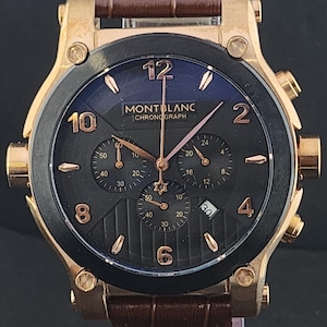 Puede incluir: Un reloj cronógrafo Montblanc con caja en tono oro rosa, esfera negra con números y marcadores en tono oro rosa, tres subesferas, ventana de fecha y correa de cuero marrón con un patrón texturizado de imitación cocodrilo.