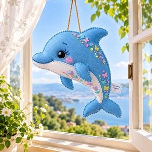 以下が含まれることがあります： 青いフェルト製のイルカの吊り下げ飾り。白い腹部、刺繍された蝶のディテール、カラフルなスパンコールが特徴です。イルカには長いまつ毛の黒い目があり、ハートのシンボルと共に「Creacione Lugme」と書かれた小さなタグが付いています。