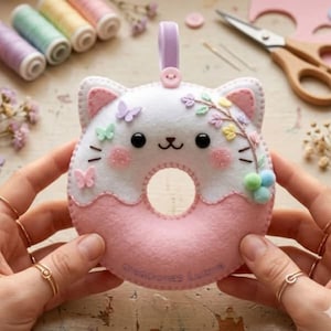 Könnte beinhalten: Ein Filzanhänger in Form eines Katzen-Donuts in Weiß und Rosa mit gestickten Gesichtszügen, kleinen Schmetterlingsapplikationen und einem Blumenzweig. Am unteren Rand steht der Sticktext Creaciones Luznie.