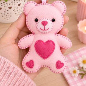 May include: A light pink felt teddy bear keychain featuring a dark pink heart appliqué on the chest and smaller dark pink hearts on each foot. The bear has black bead eyes, a stitched nose and mouth, and visible blanket stitching along the edges.