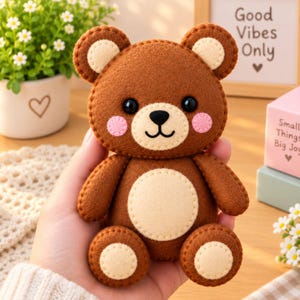 May include: A small brown felt teddy bear with a cream-colored belly, ear patches, and foot pads. The bear features black bead eyes, a stitched black nose and mouth, and pink circular felt cheeks. The edges are finished with visible brown stitching.