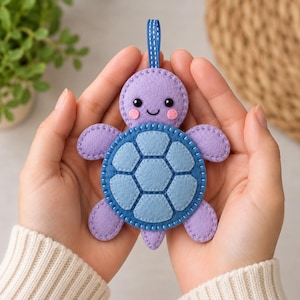 May include: A felt turtle ornament featuring a light purple head with pink cheeks and black eyes, and a blue shell with a hexagonal pattern. The turtle has four purple flippers and a small blue hanging loop at the top, all stitched with white thread.