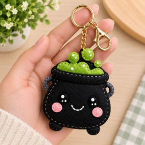 May include: A black felt keychain shaped like a cauldron with a smiling face, pink cheeks, and bright green bubbling liquid at the top. The cauldron features white stitching, grey side handles, and is attached to a gold-toned metal ring and clasp.