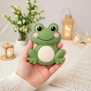 May include: A green felt frog ornament featuring black eyes, pink circular cheeks, and a cream-colored belly patch. The frog has stitched edges and a simple embroidered smile, crafted from soft felt material in a flat, layered design.