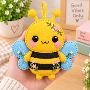 May include: A yellow and black felt bee ornament with light blue wings, featuring embroidered pink and white flowers, two black bead eyes, pink cheeks, and a small yellow hanging loop at the top. The bee has a rounded body with two black stripes.